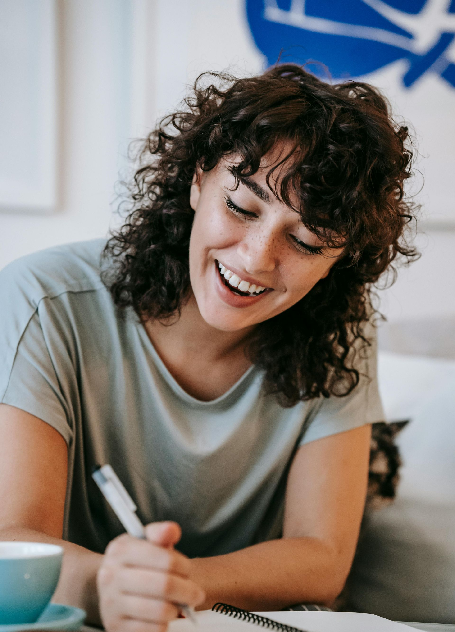 Woman with curly hair, smiling while writing in a notebook; light green top, pen in hand.