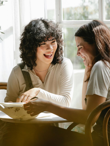 Two people laugh while seated at a table near a window. One holds a bowl, the other smiles.