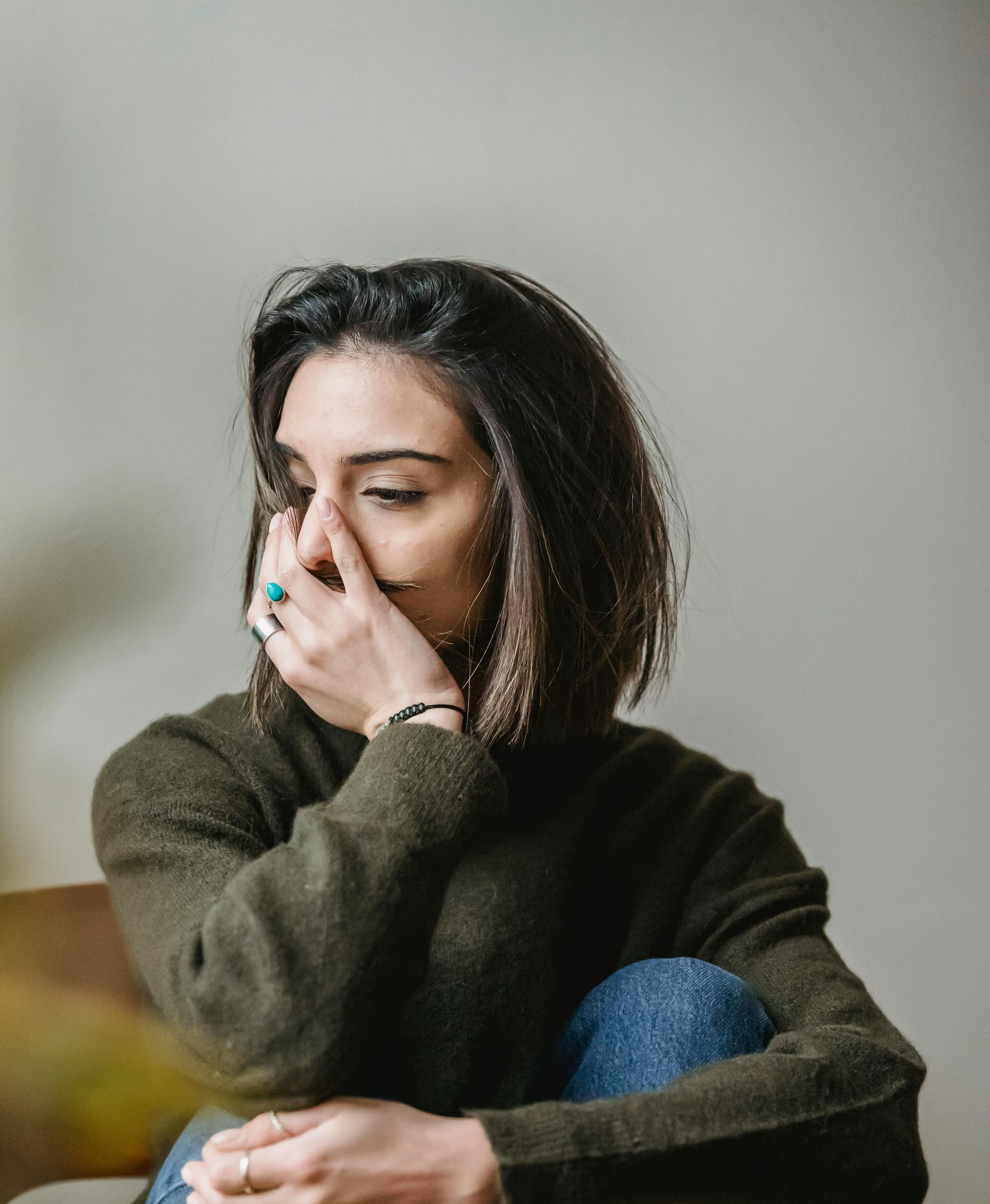 Woman with hand covering her face, looking down, wearing a green sweater and blue jeans.
