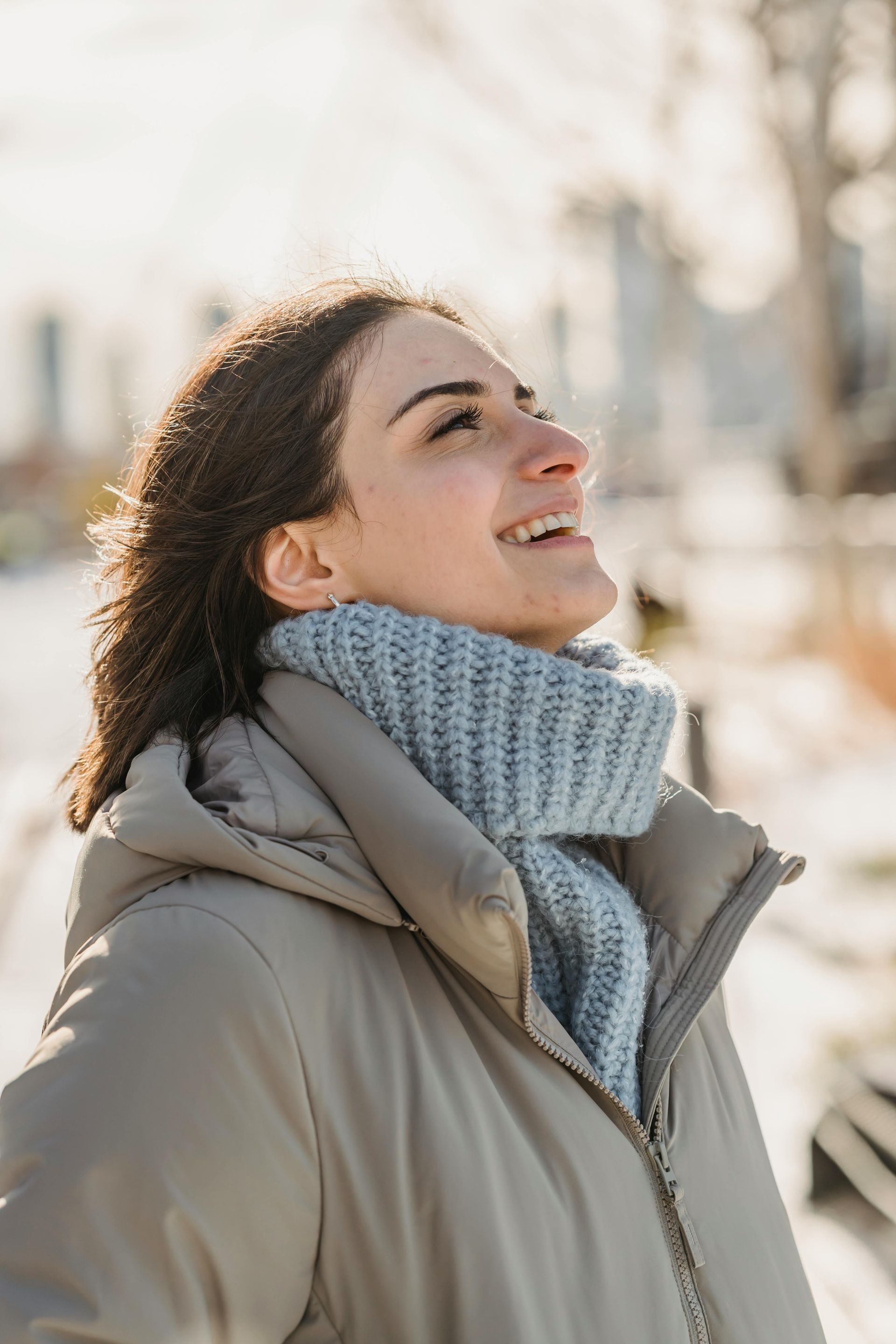 Woman smiling, looking up at the sunlight, wearing a light-colored puffer jacket and blue scarf outdoors.