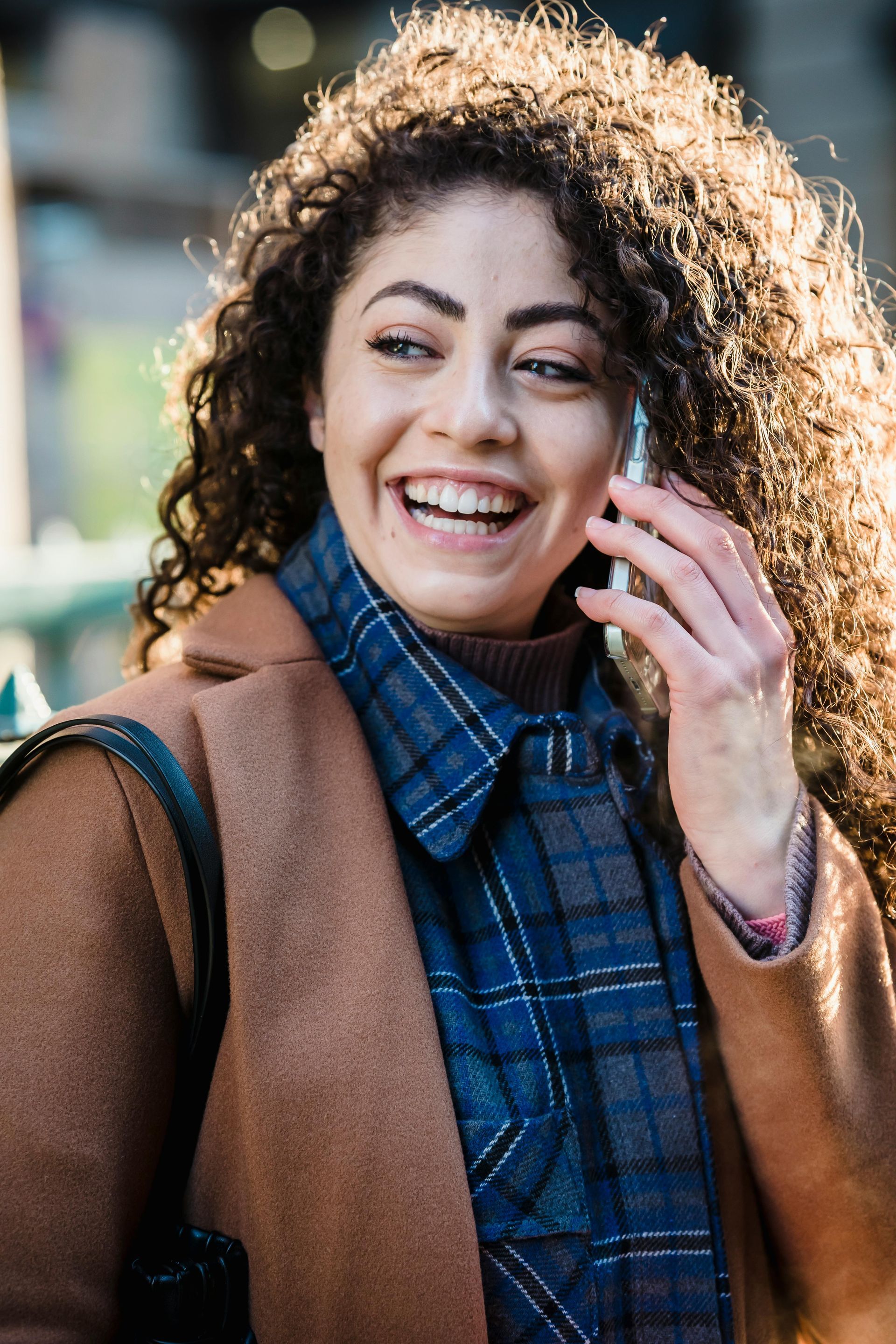 Woman with curly hair smiles while talking on a phone, wearing a brown coat and blue plaid shirt.