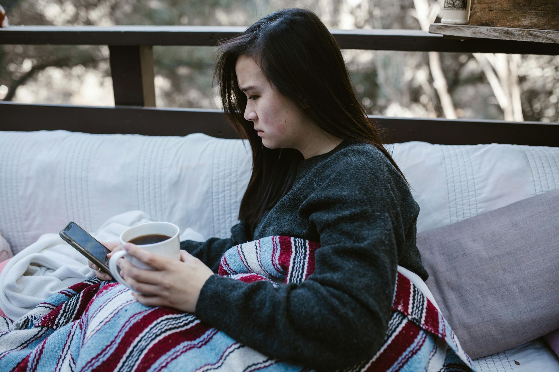 Woman wrapped in blanket on a porch, holding a mug and a phone, looking down.