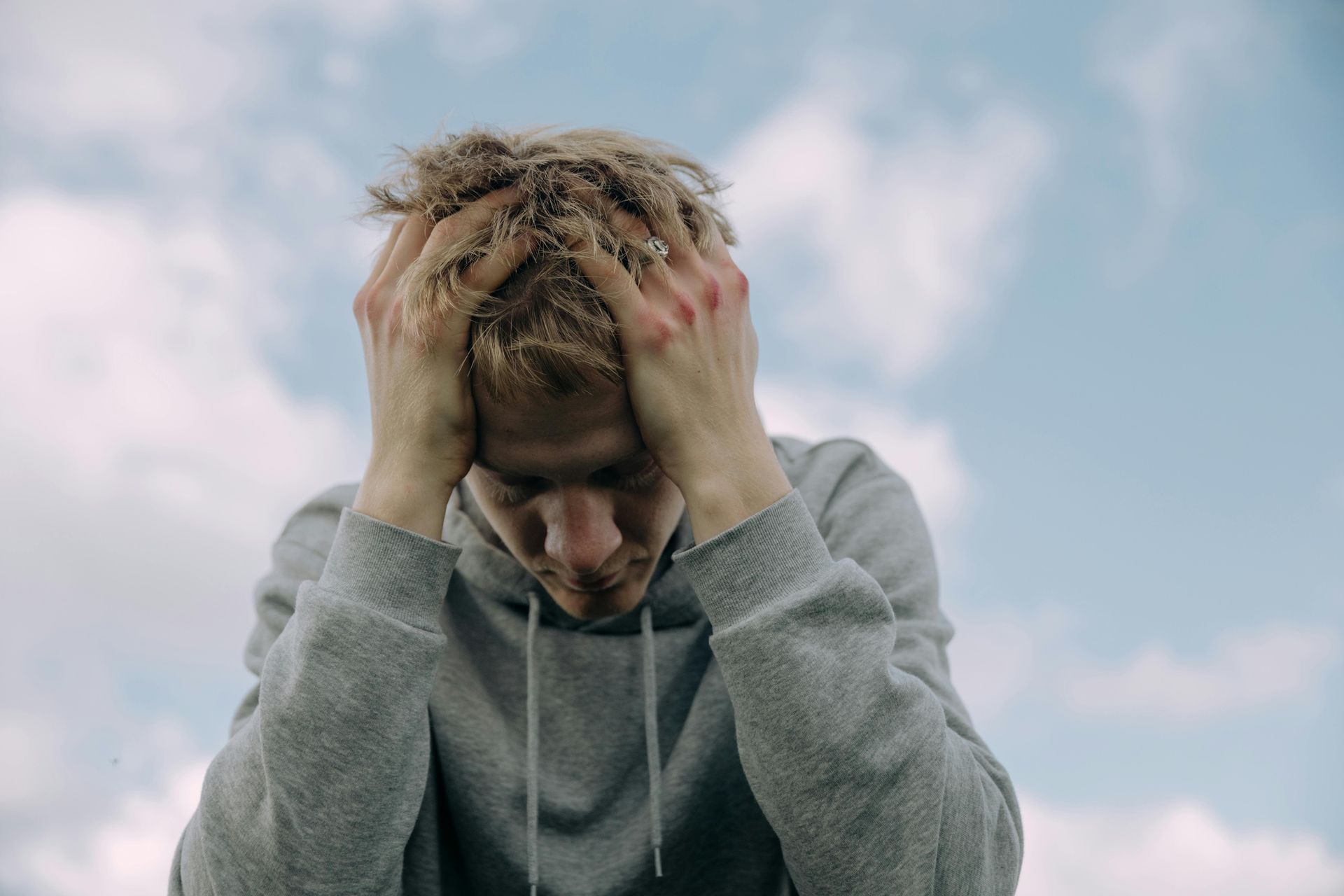 Person with head in hands, distressed expression, gray hoodie, outdoors with cloudy sky.