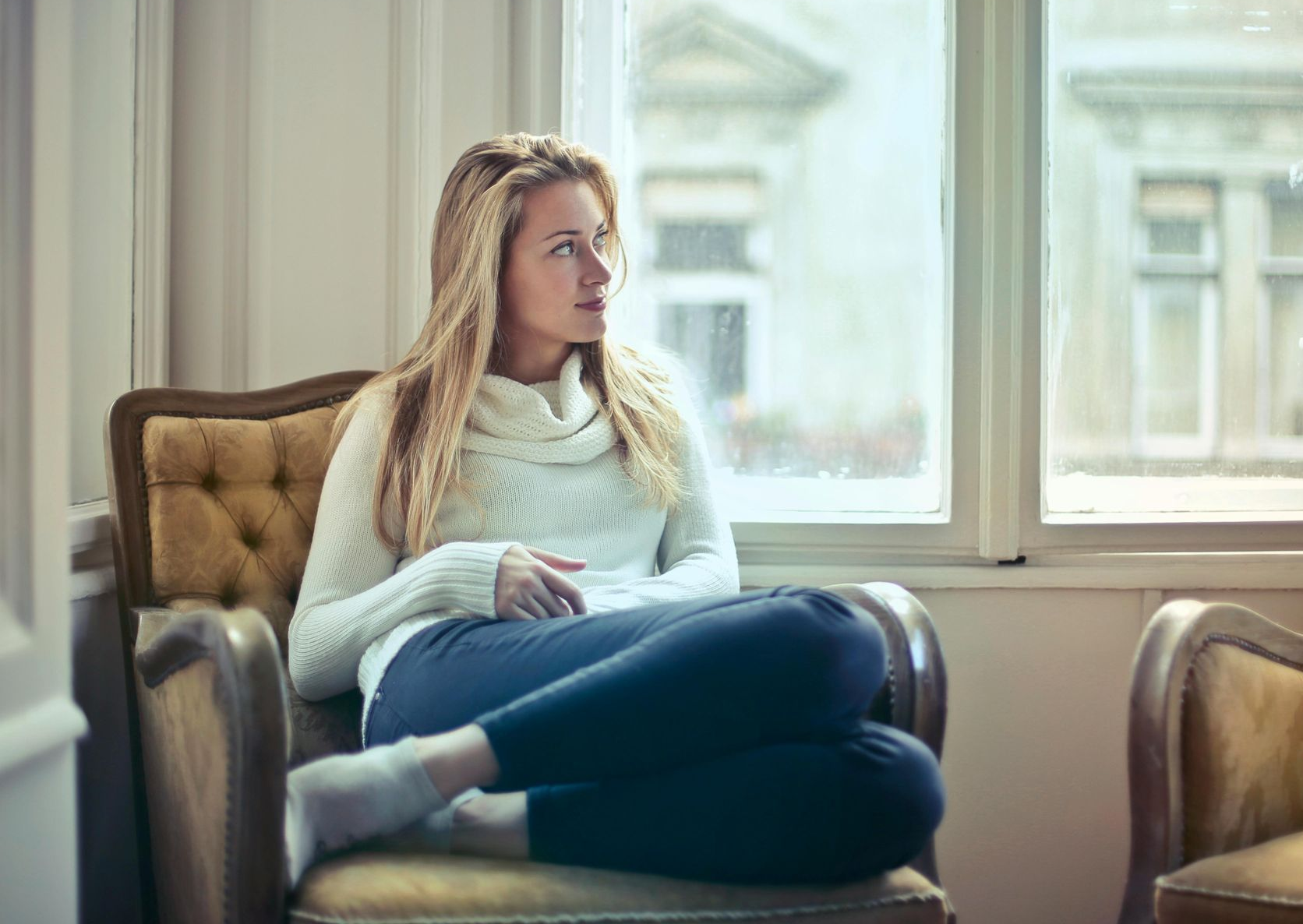 Woman in a white sweater and blue pants looks out a window from a yellow armchair.