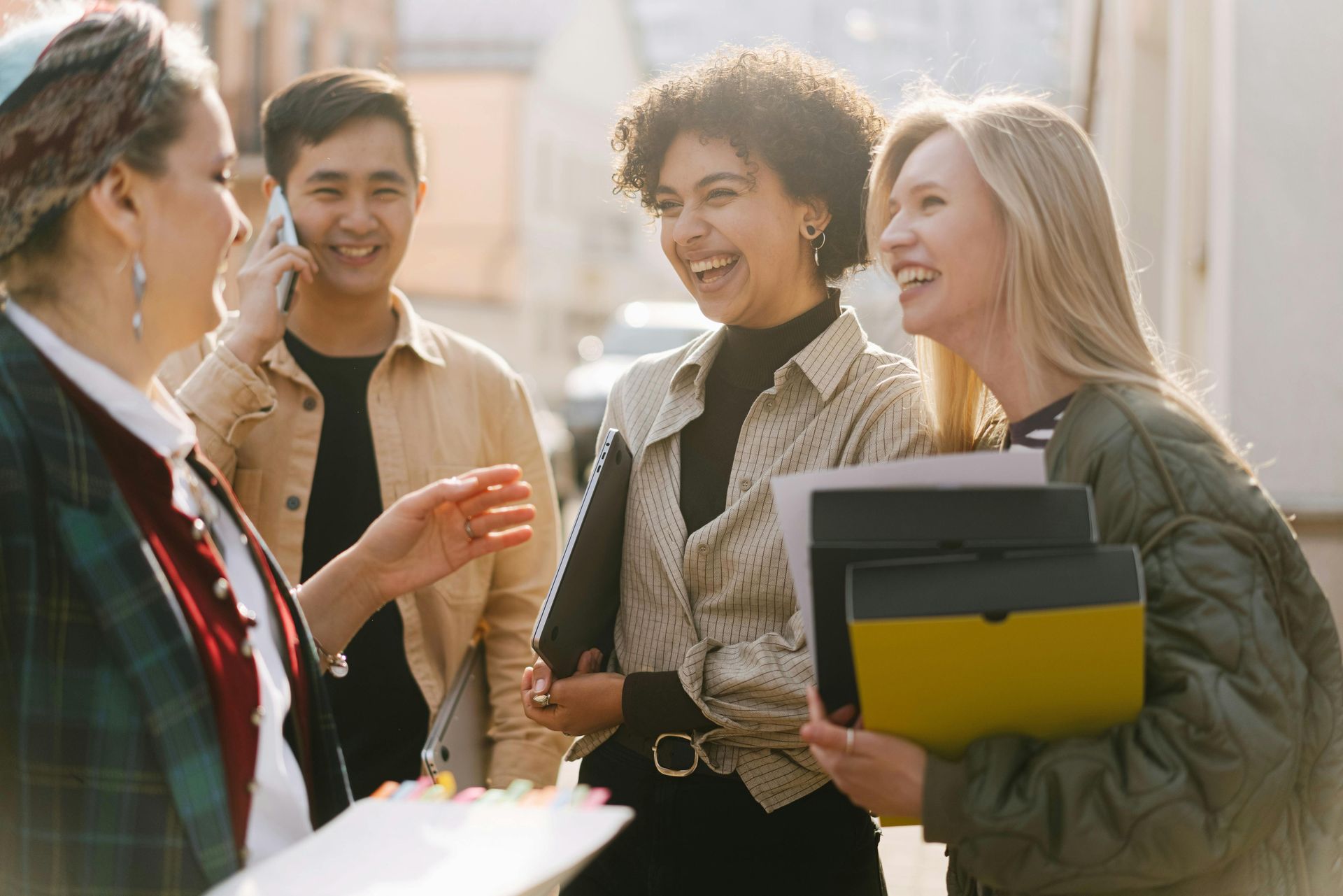 Group of people laughing outdoors; two holding folders, one on phone.