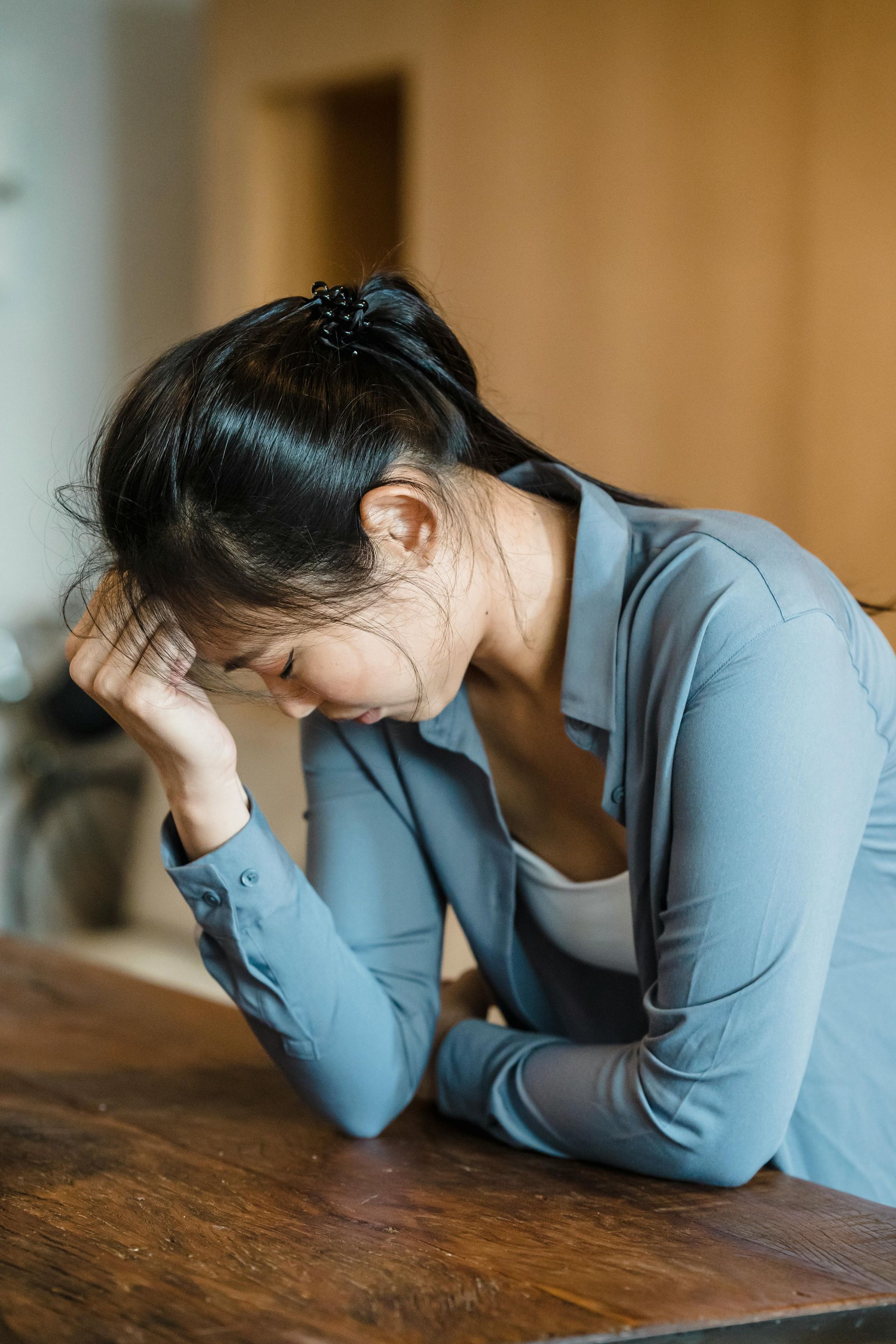 Woman resting head on hand at wooden table, looking down, appearing stressed.