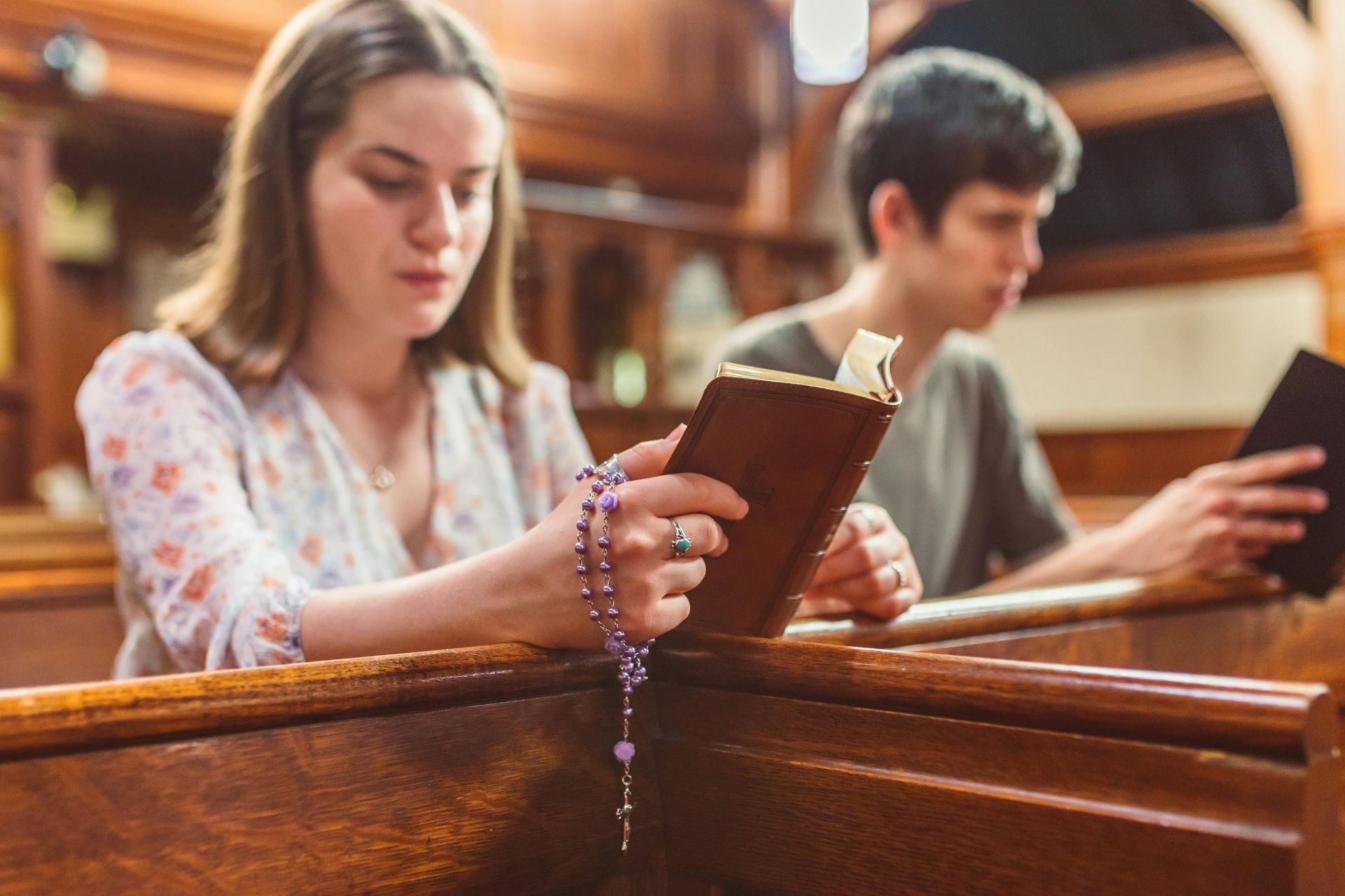 Woman and man reading books in a church pew. Woman holds rosary beads.