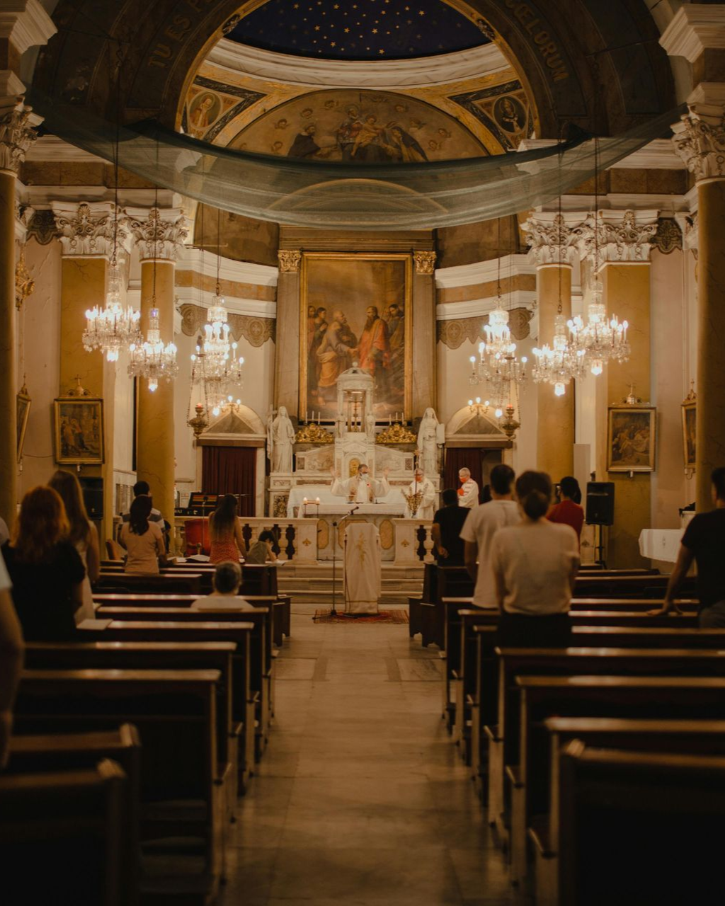 Interior of a church during a service. People seated in pews face the altar where a priest is speaking.