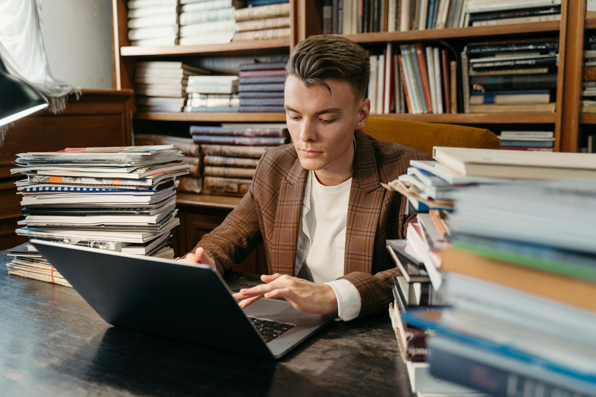 A person in a brown blazer types on a laptop at a desk, surrounded by stacks of books and bookshelves.