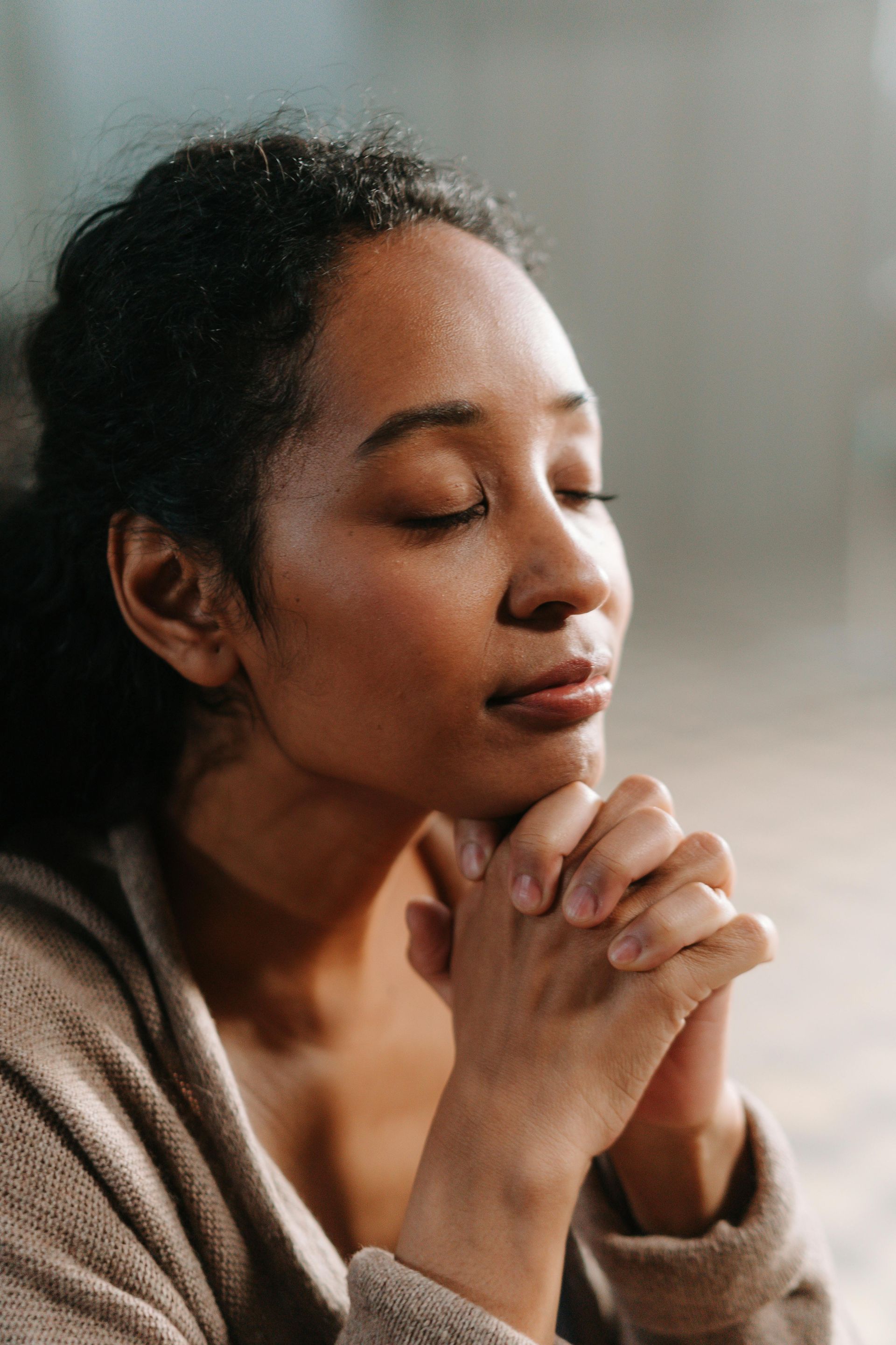 Woman with eyes closed, hands clasped, in prayer or contemplation; neutral tones.