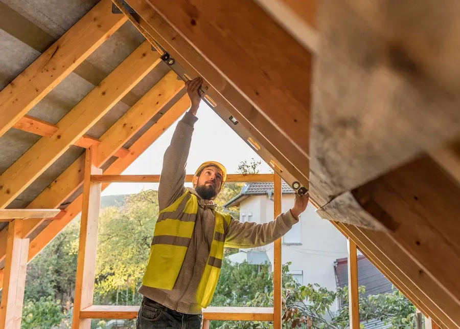 Worker in a yellow safety vest inspects attic roof framing during construction.