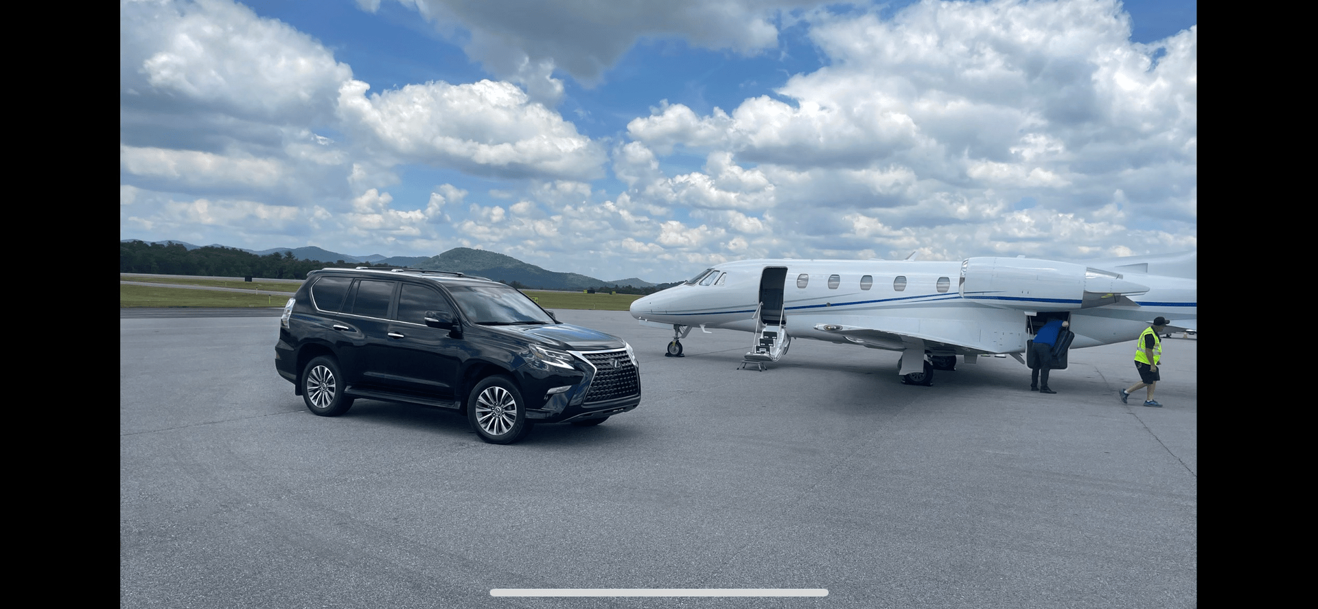 Black SUV next to a private jet on an airfield with a cloudy sky.