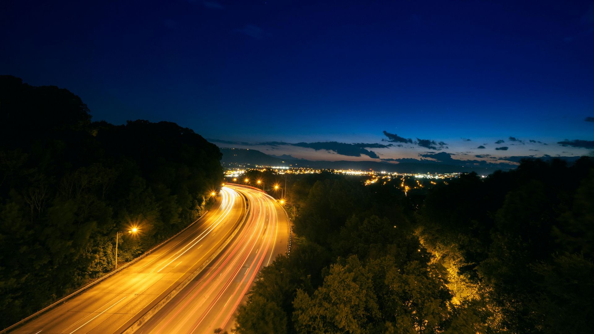 Highway at dusk with illuminated car trails, bordered by dark trees, and city lights in the distance.