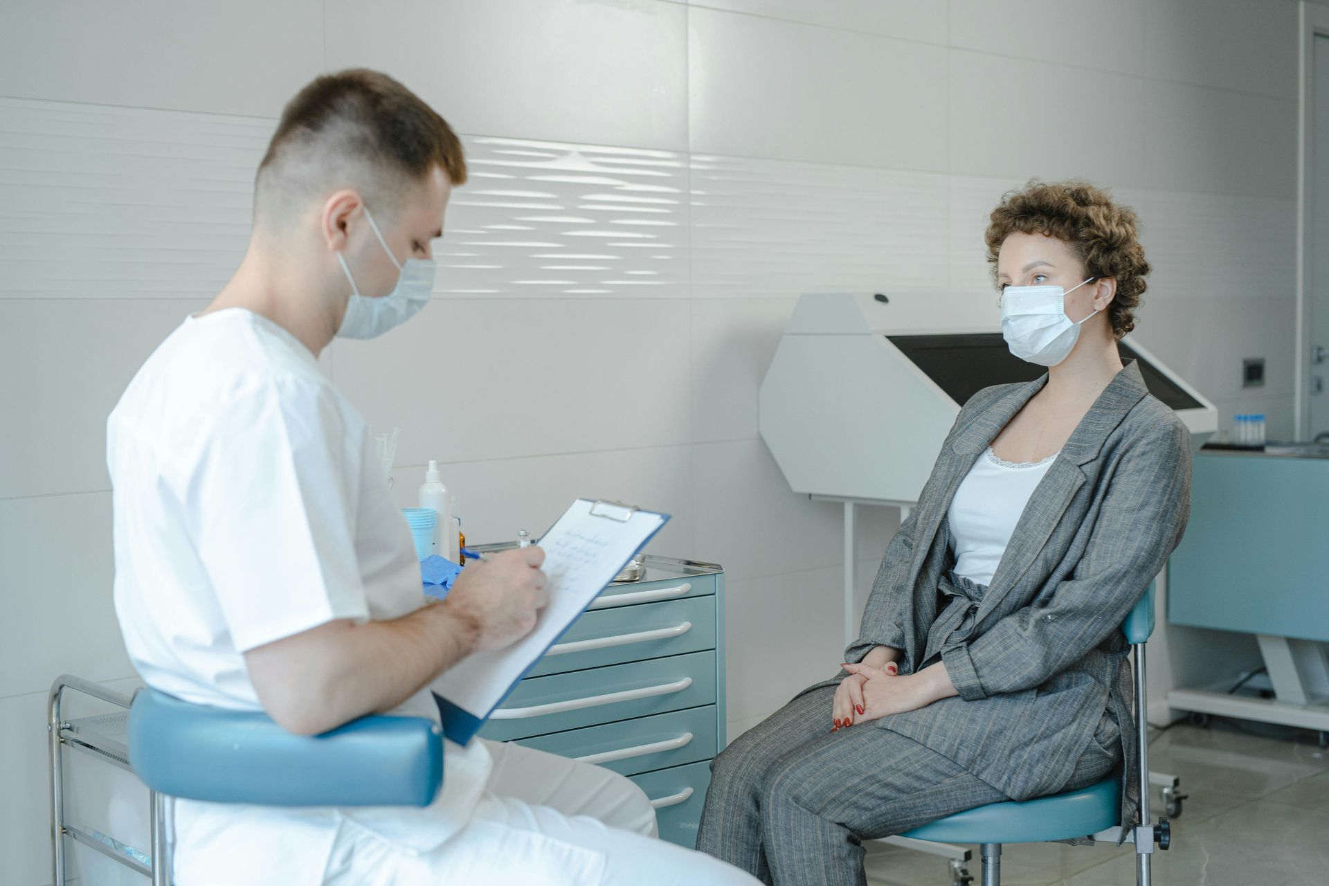 Doctor in mask taking notes while talking to patient in mask, seated in a medical setting.