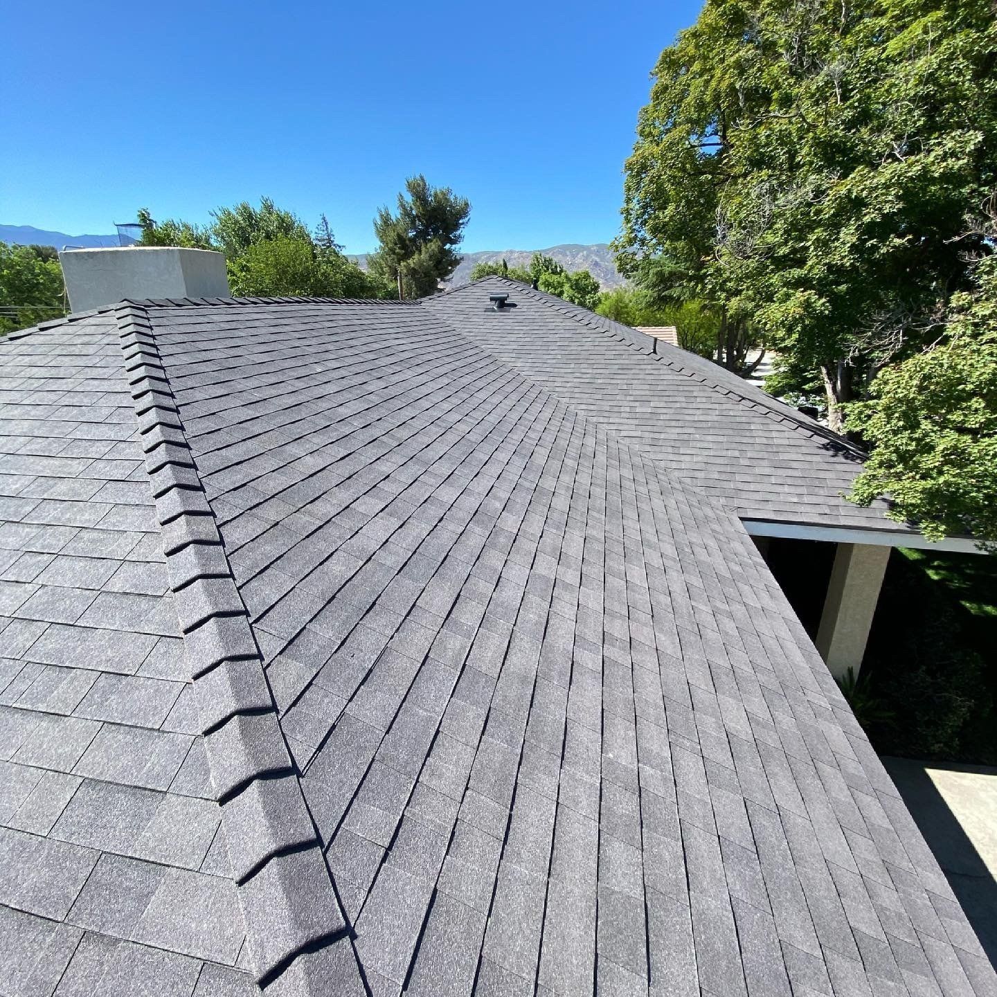 Gray asphalt shingle roof on a house under a blue sky, surrounded by green trees