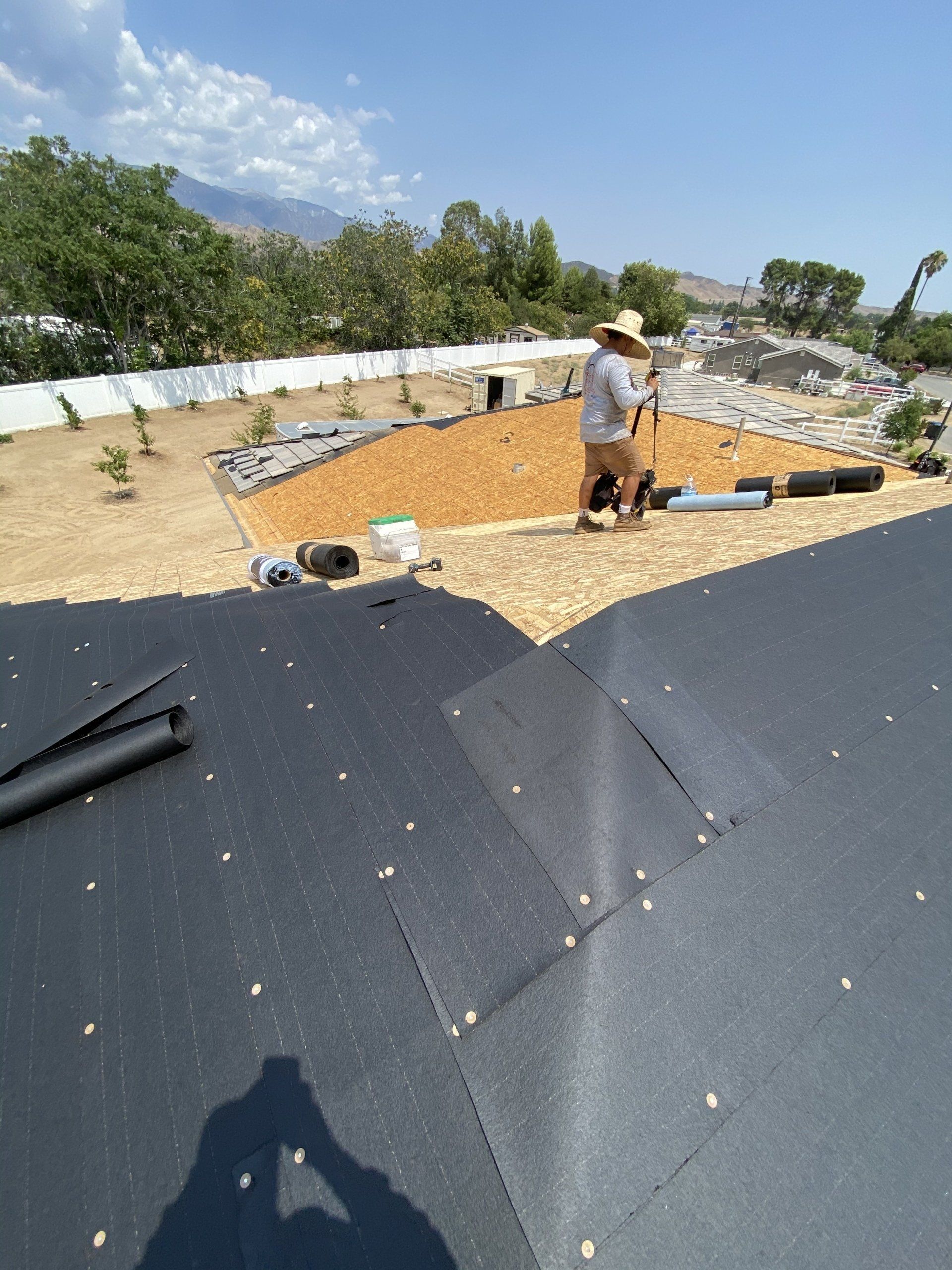 Man on roof with landscaping in progress, tan gravel, black underlayment, blue sky, trees, mountains in background.