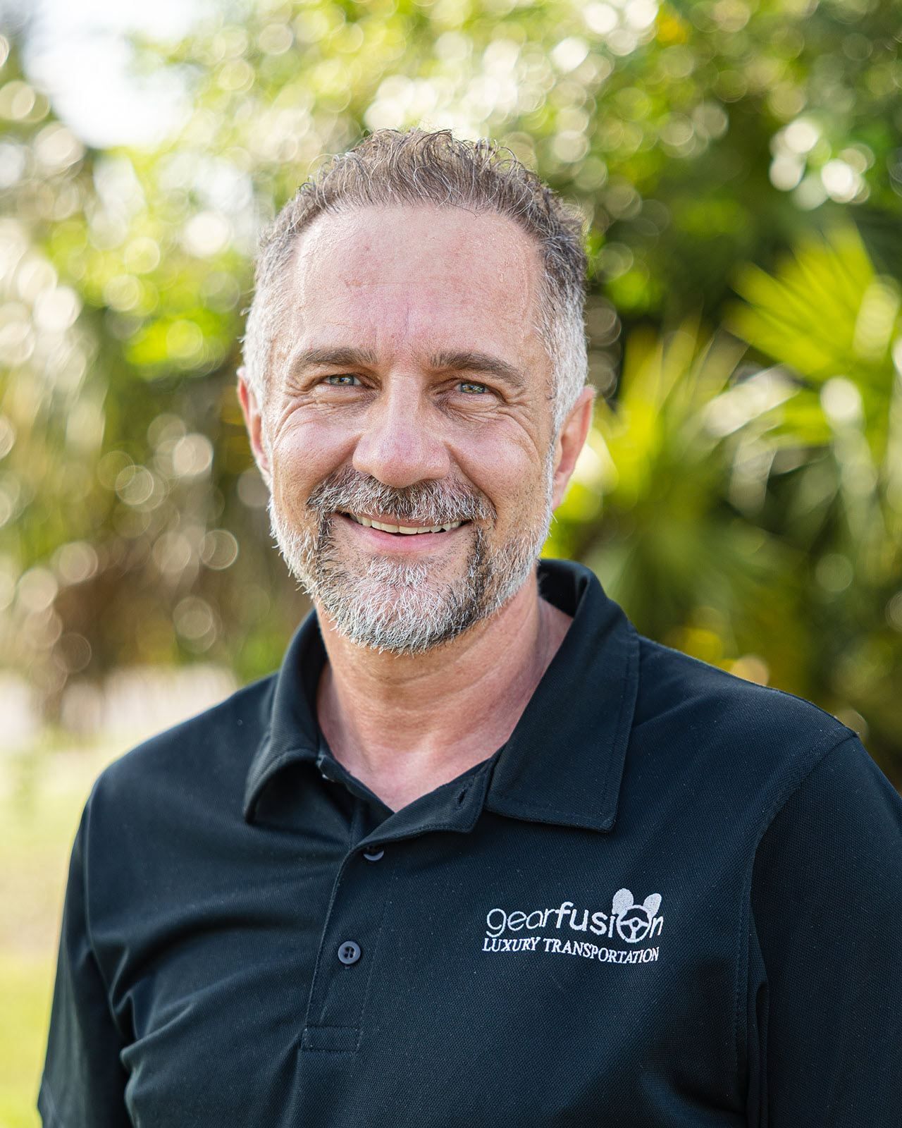 A man with a beard is wearing a black polo shirt and smiling.