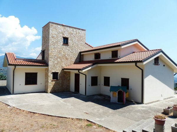 Modern stucco house with stone tower, red-tiled roofs, and a sunny concrete driveway