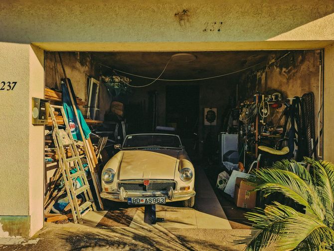 Vintage car parked in a cluttered garage with tools, ladders, and plants around it