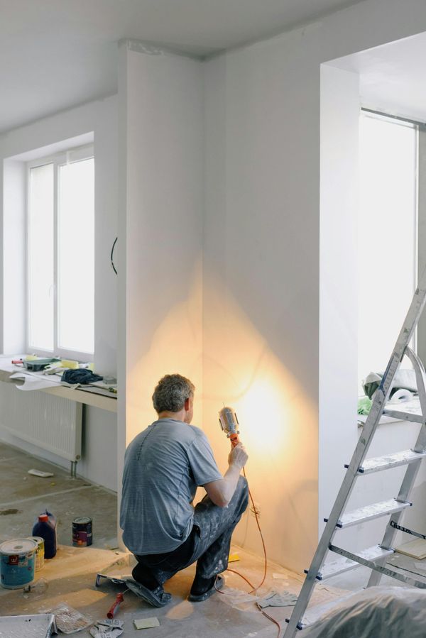 Person kneels painting a white wall corner while using a flashlight; ladder and supplies nearby.