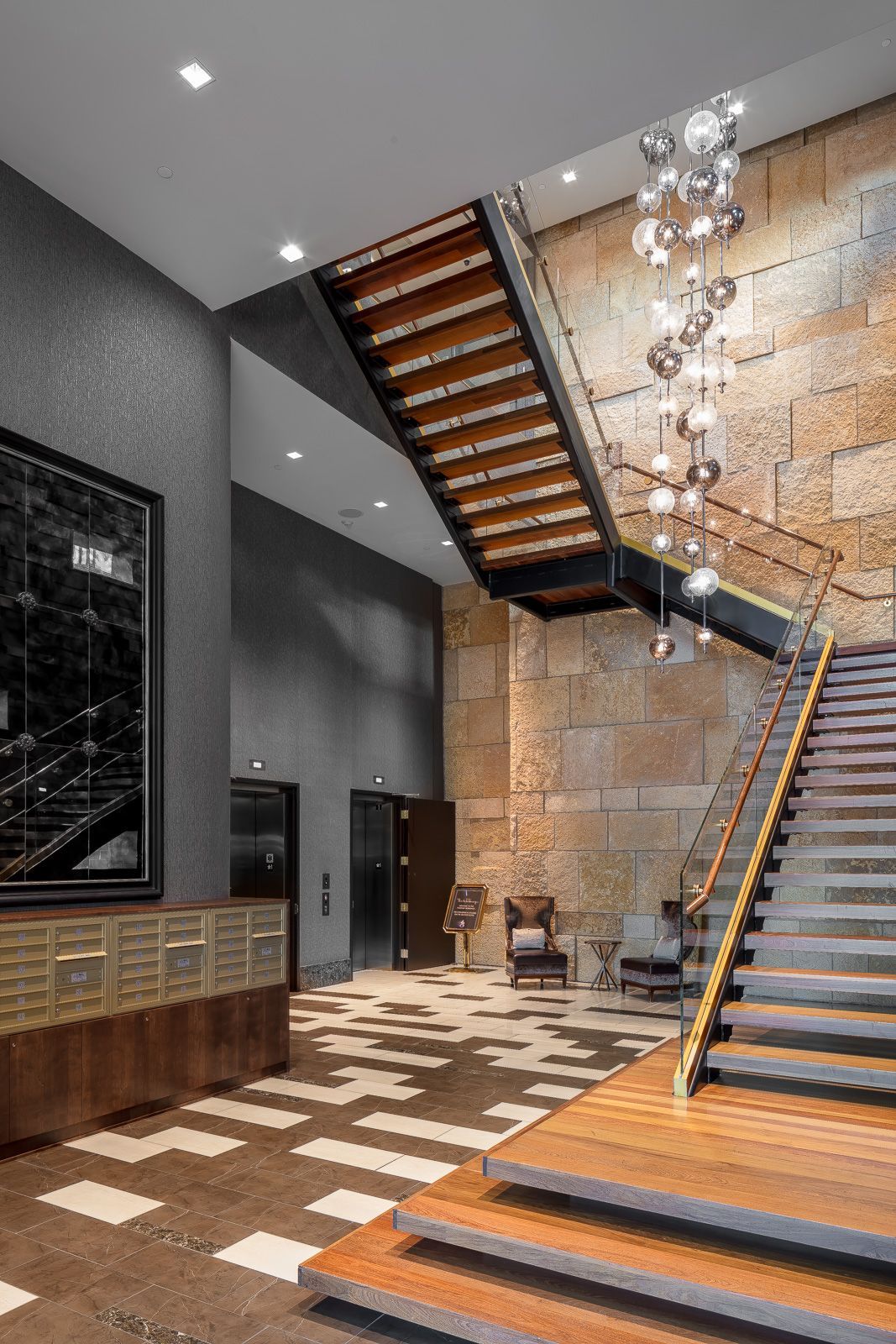 Modern apartment lobby with curved wooden staircase, stone wall, chandelier, and elevators.
