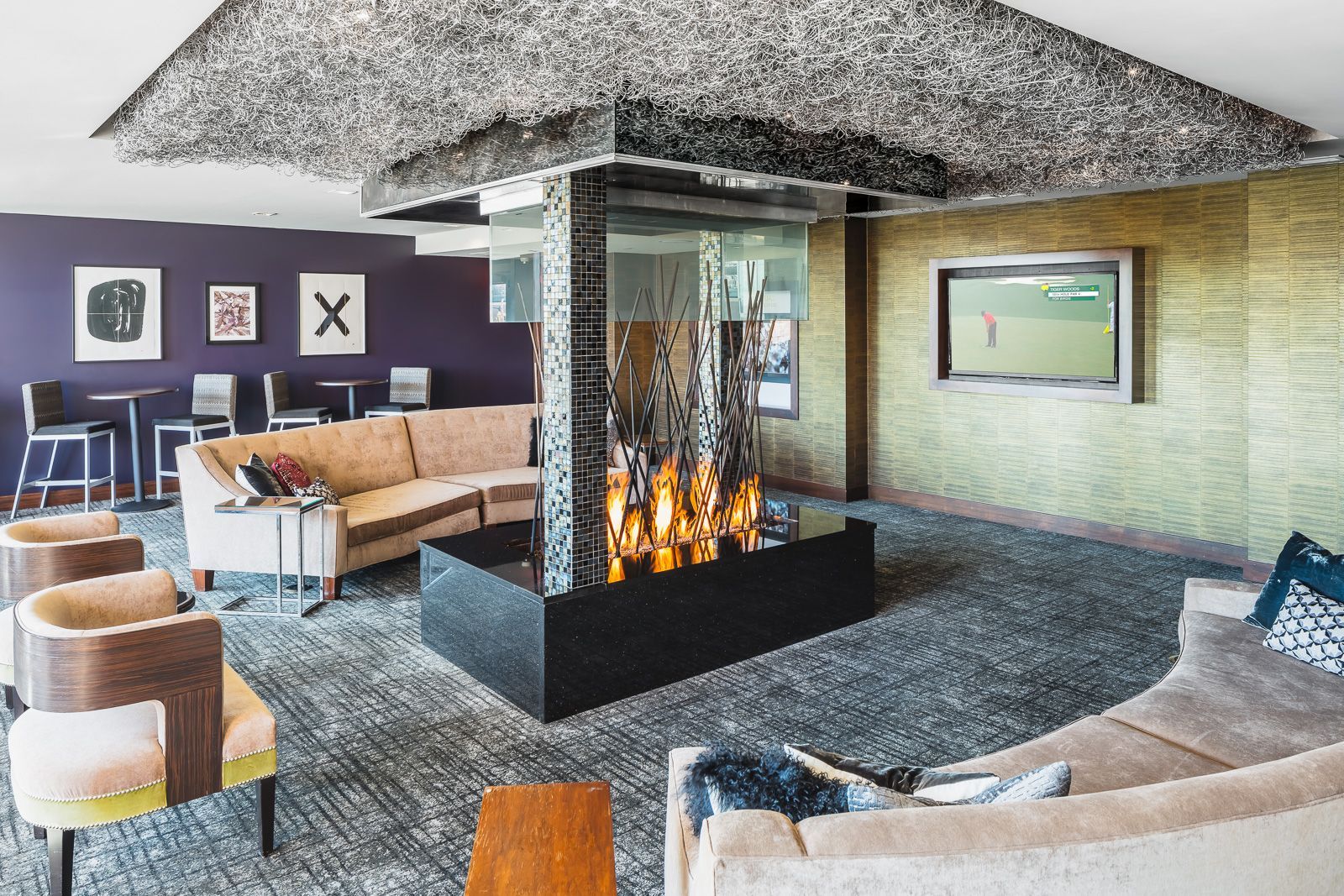 Lounge lobby with beige sectional seating, a central fire feature, purple accent wall, and a wall-mounted TV.