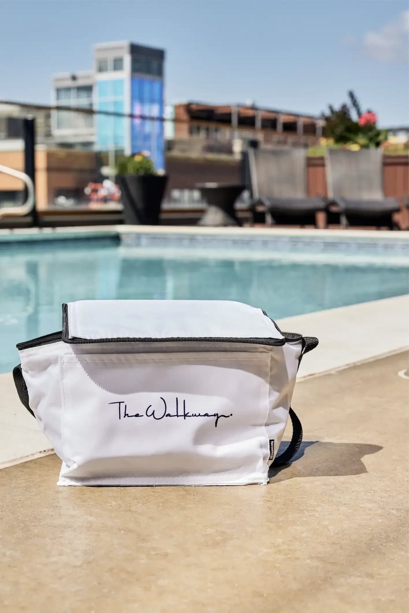 White bag with a black zipper sits on a pool deck; lounge chairs and a modern building are visible in the background.