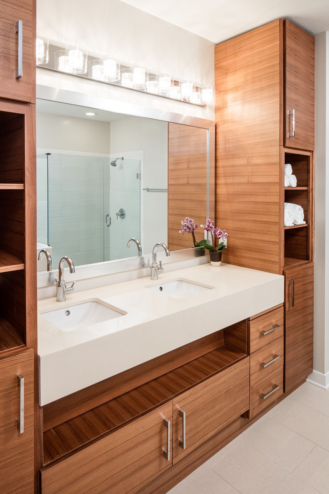 Double-sink bathroom vanity with white countertop, wood cabinetry, and a glass-enclosed shower in the background.