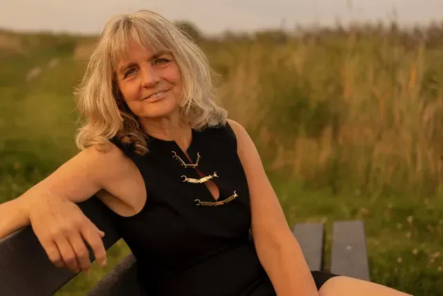 Een vrouw met blond haar, gekleed in een zwarte jurk, lacht op een bankje buiten in een veld. Business coach uit Friesland