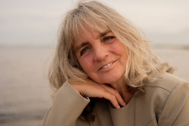 Een vrouw met blond haar lacht, haar arm rustend op haar jas. Strandachtergrond, zacht licht. Business coach