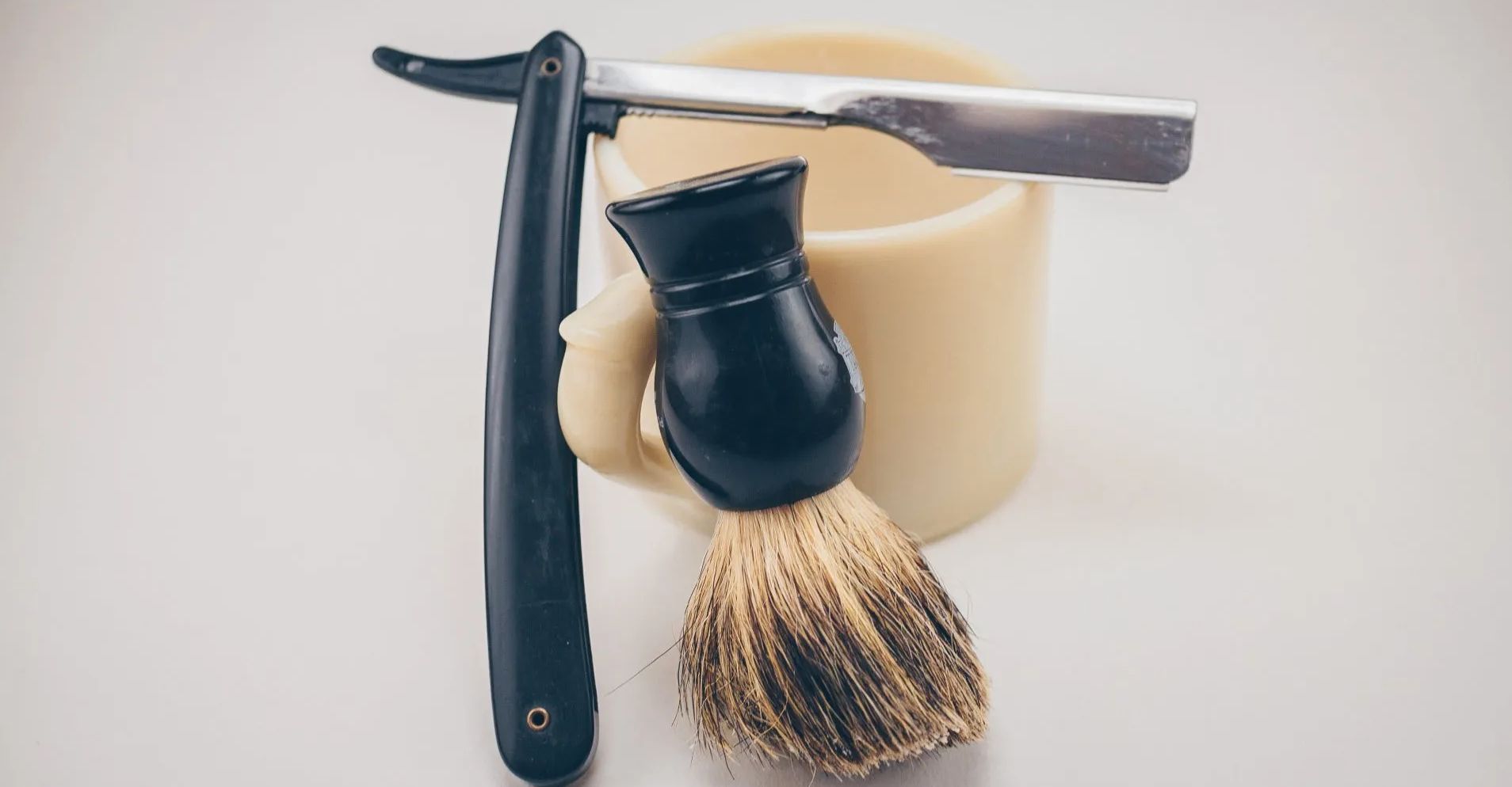 Straight razor, shaving brush, and mug on a white surface, ready for shaving.