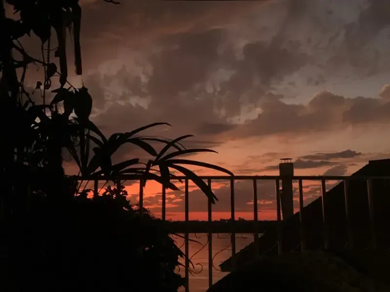 Pôr do sol sobre a água com a silhueta da cerca, plantas e nuvens dramáticas em tons de laranja e cinza.