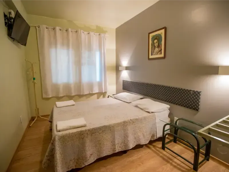 Quarto com cama de casal, mesas de cabeceira, janela e luminárias de parede. Piso marrom claro e paredes cinza.