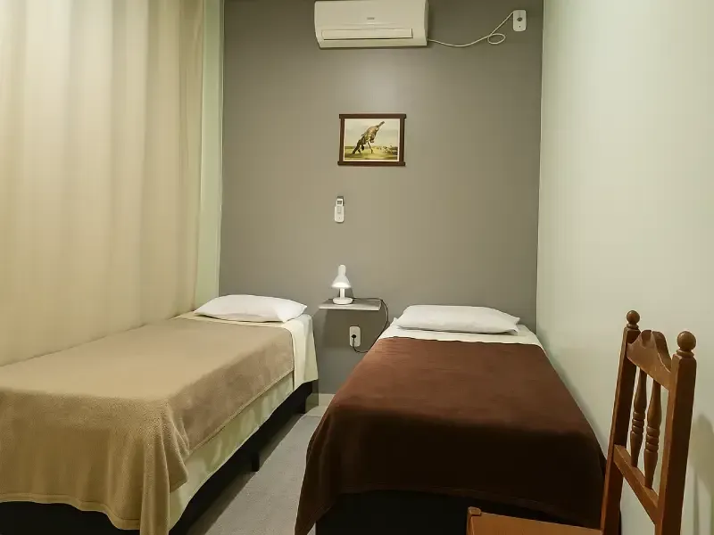 Quarto com duas camas de solteiro. Uma com cobertor bege, a outra com cobertor marrom. Paredes cinza e branca.