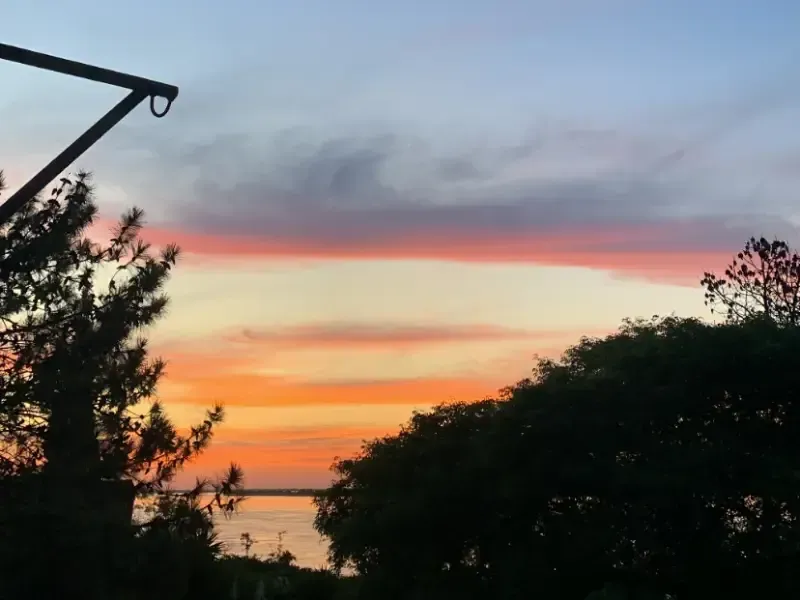 Pôr do sol sobre a água com tons de laranja e rosa, emoldurado por árvores em silhueta e uma viga de sustentação preta.