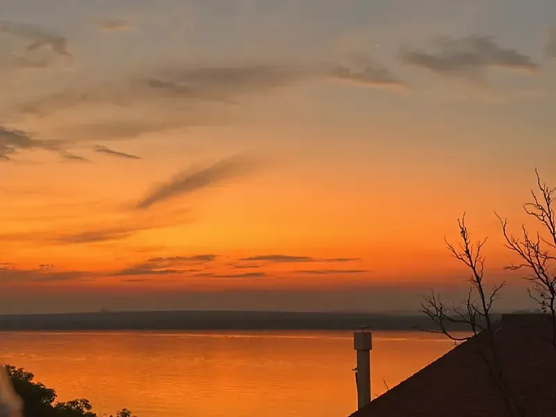 Pôr do sol alaranjado sobre águas calmas; silhuetas de árvores e telhados em primeiro plano.