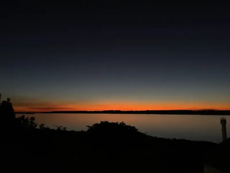 Pôr do sol sobre um corpo d'água, com a silhueta escura de terra e árvores em primeiro plano, céu alaranjado e azul.