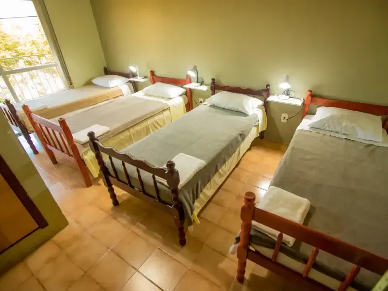 Quarto com quatro camas de solteiro, cada uma com um abajur e uma toalha aos pés da cama. Paredes verdes.