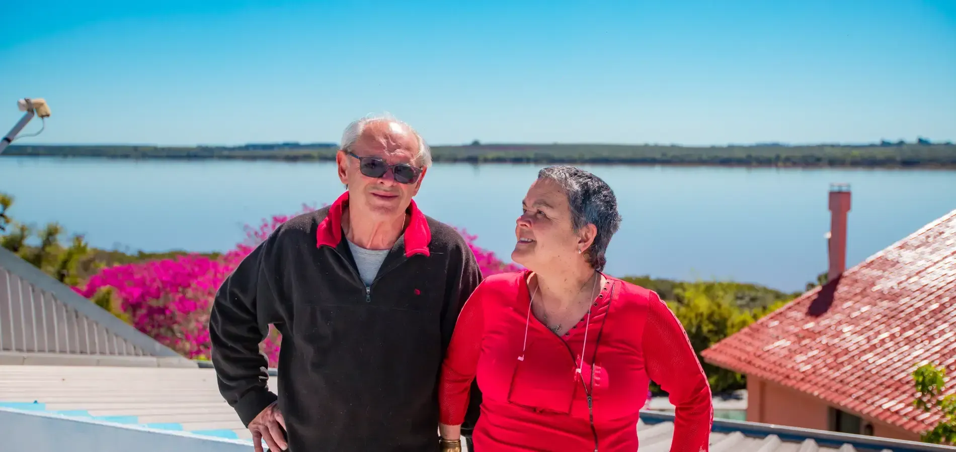 Duas pessoas em um terraço com vista para o lago; a mulher sorri para o homem que usa óculos de sol e um suéter preto.