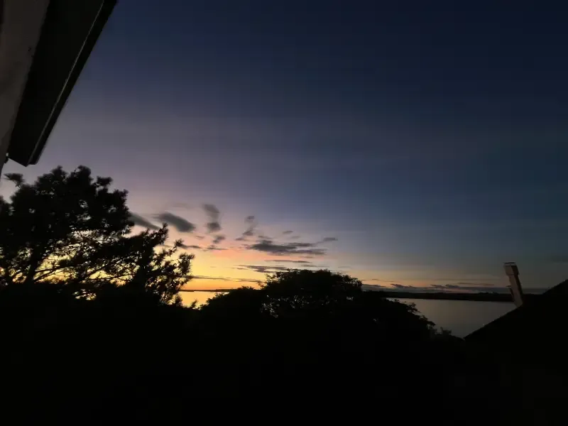 Silhuetas de árvores e edifícios emolduram um pôr do sol colorido sobre a água, com tons de laranja, amarelo e azul.