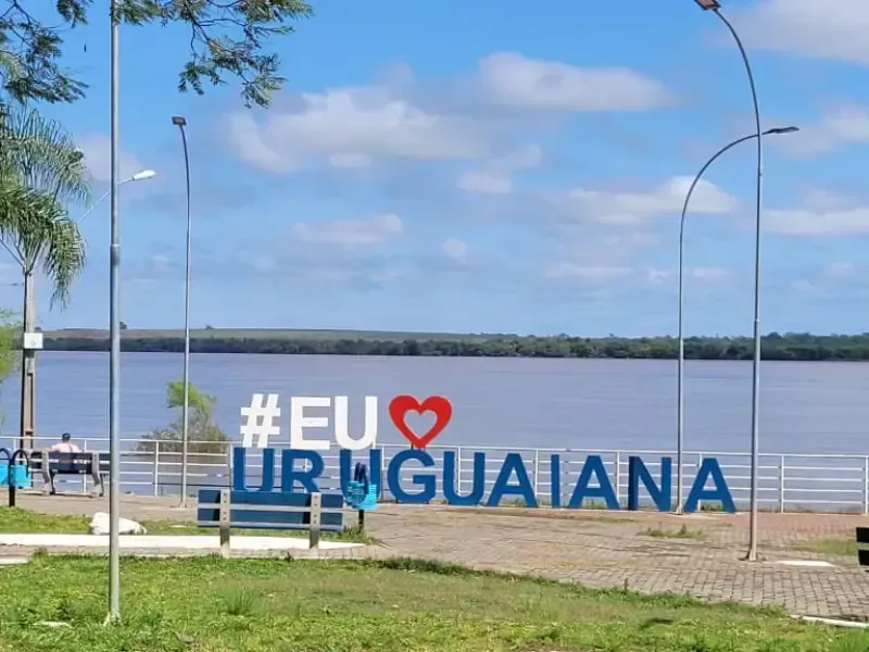Placa com a hashtag #EU❤️URUGUAIANA com vista para um rio, com letras azuis e brancas em uma área gramada.