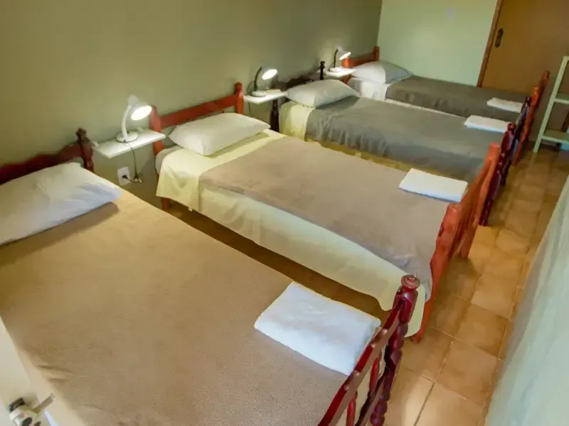 Quatro camas com lençóis brancos e cobertores bege em um quarto com piso de azulejo. Cada cama possui uma toalha branca.