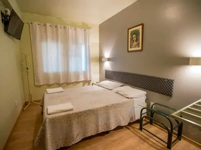 Quarto com cama de casal, parede cinza, janela e TV.