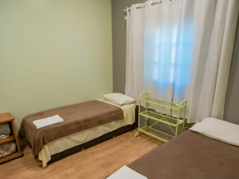 Duas camas em um quarto com paredes verde-claras, uma janela e uma pequena estante.