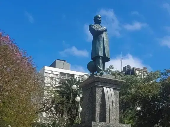 Estátua de um homem de casaco, com as mãos juntas em oração, sobre um pedestal de pedra em um parque, com prédios ao fundo.