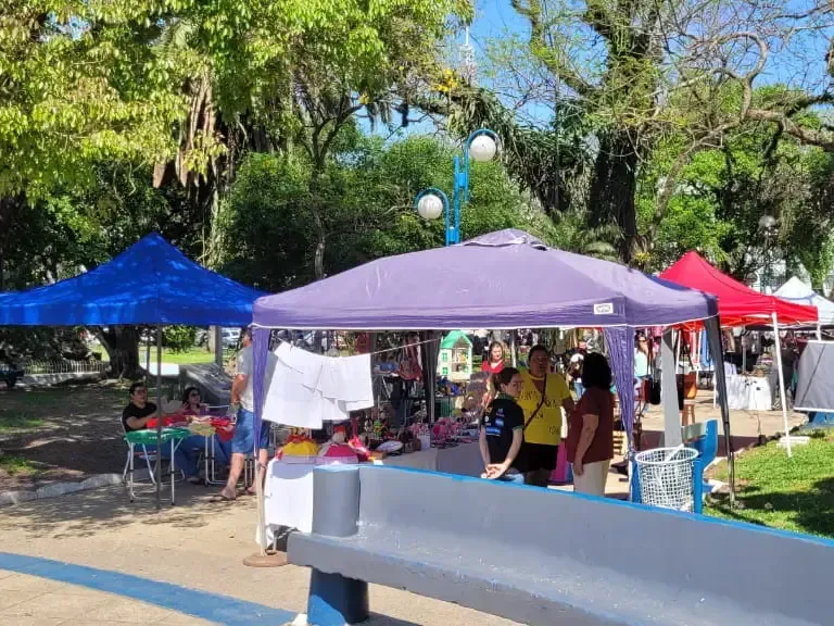 Vendedores ambulantes sob tendas coloridas; compradores observam os artesanatos.