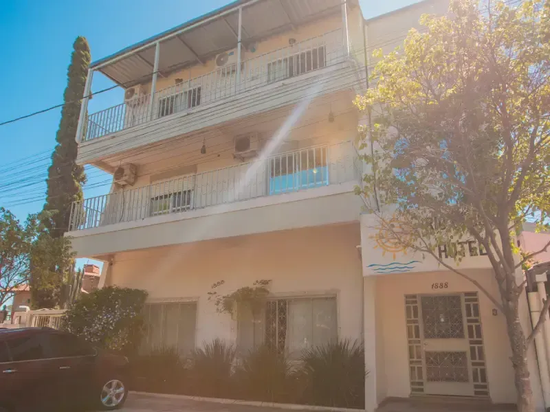 Prédio de hotel com varandas, fachada bege e uma árvore alta em um dia ensolarado.