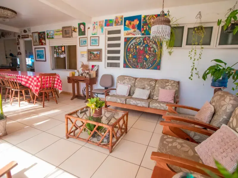 Espaço de convivência externo banhado de sol, com paredes repletas de arte, um sofá, cadeiras, uma mesa de jantar e plantas.