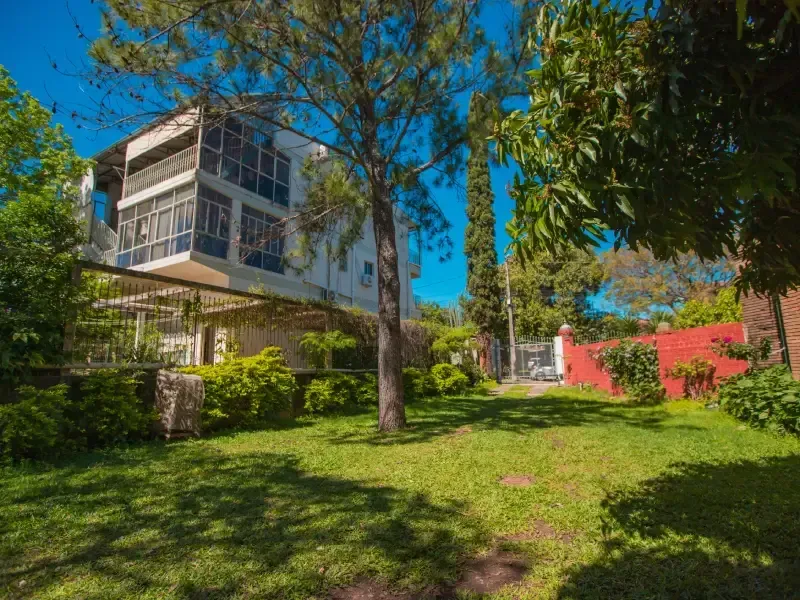 Casa branca de dois andares com gramado verde, árvores e muro de tijolos vermelhos no quintal, em um dia ensolarado.