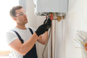 A worker wearing safety glasses and black gloves uses a wrench to install plumbing components on a wall-mounted boiler.